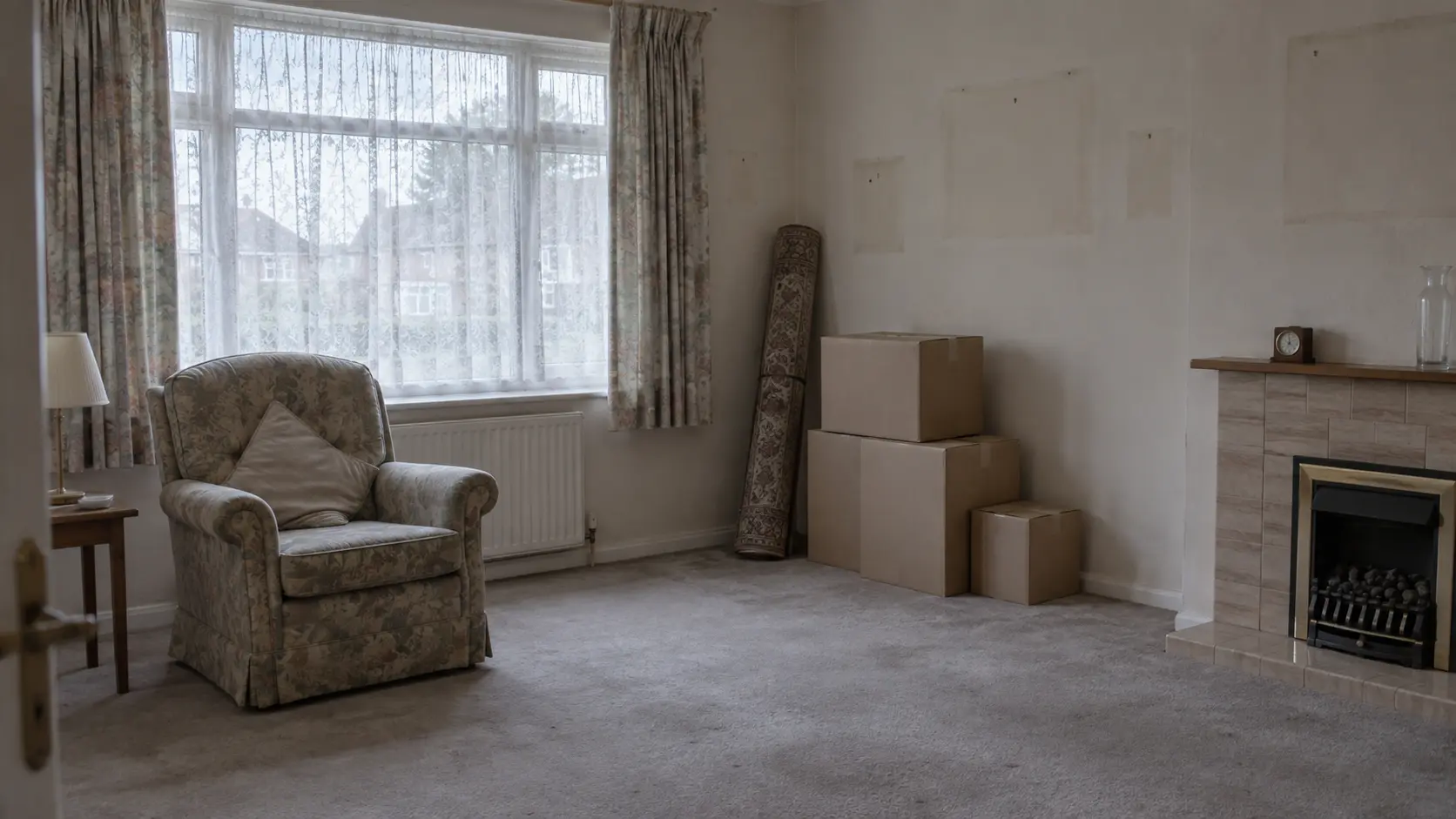 A quiet living room mid-clearance, with an armchair, rolled-up rug and packed boxes left beside an empty fireplace